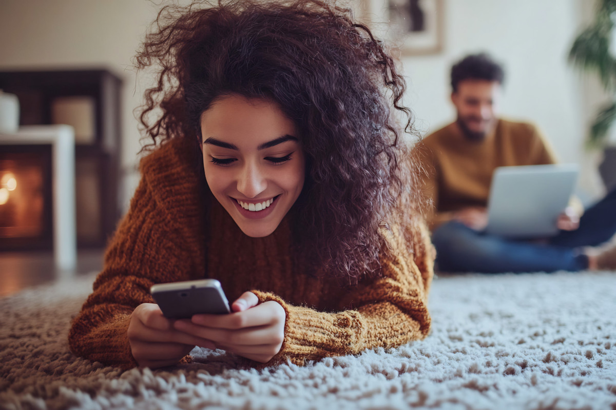 Frau liegt lächelnd auf dem Boden und blickt in ihr Smartphone, unscharf im Hintergrund sitzt ein Mann mit Laptop